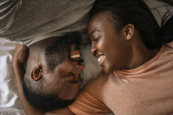 Young African American couple lovingly smiling at each other while lying face to face in bed together in the morning - Фото, зображення