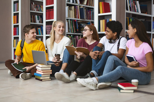 Συμμαθητές κάθονται στο πάτωμα στη βιβλιοθήκη, συζητώντας ενώ σπουδάζουν - Φωτογραφία, εικόνα