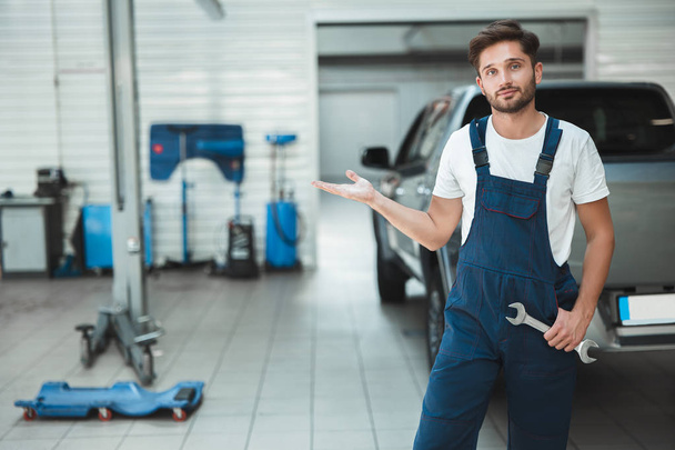 young handsome mechanic wearing uniform with spanner in his hand showing modern equipment in car service center - Foto, imagen