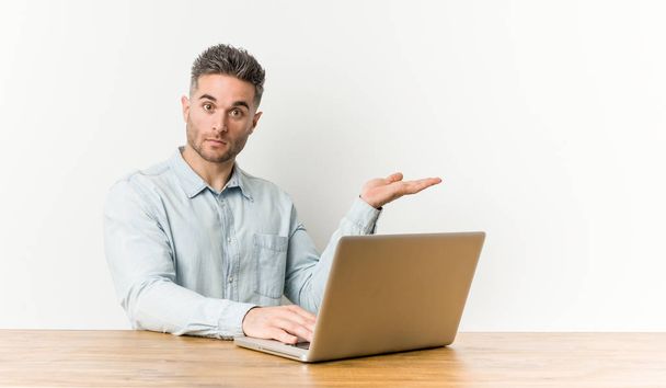 junger gutaussehender Mann, der mit seinem Laptop arbeitet, einen Kopierplatz auf einer Handfläche zeigt und eine andere Hand auf der Taille hält. - Foto, Bild