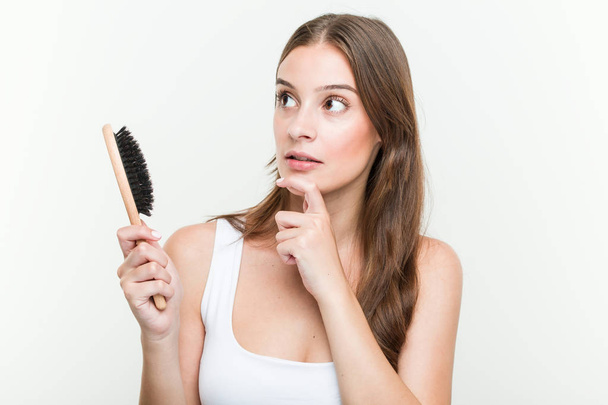 Νέα καυκάσια γυναίκα κρατώντας μια βούρτσα μαλλιών κοιτάζοντας πλάγια με αμφίβολη και επιφυλακτική έκφραση. - Φωτογραφία, εικόνα