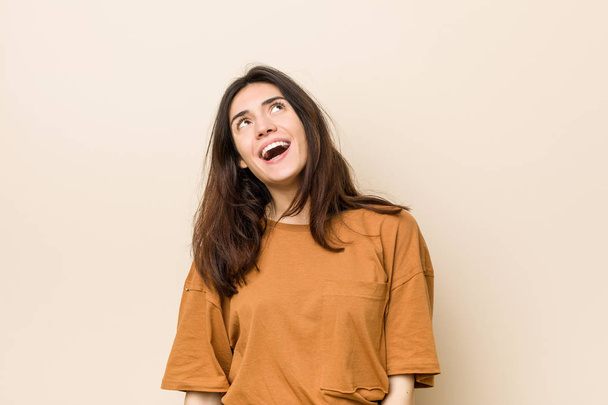 Young brunette woman against a beige background relaxed and happy laughing, neck stretched showing teeth. - Фото, зображення