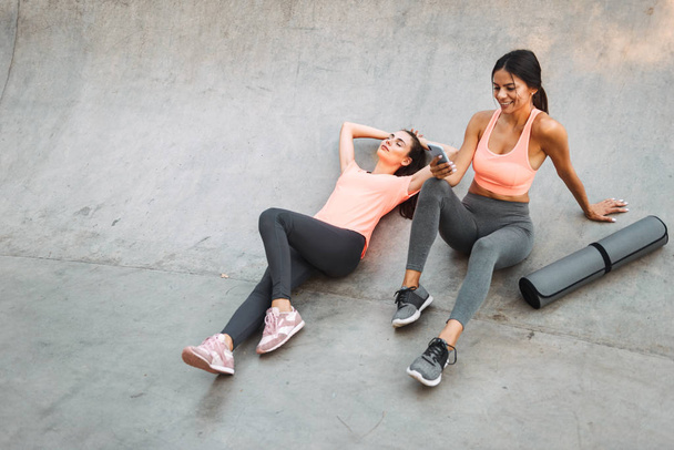Image of young fitness women resting and holding cellphone while sitting on concrete sports ground - Valokuva, kuva
