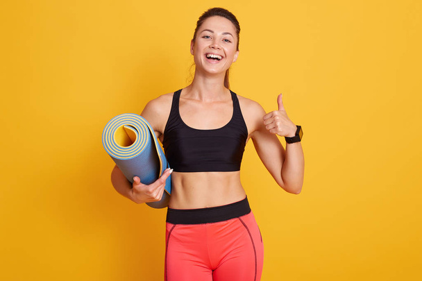 Image of slim beautiful woman ready for workout with yoga mat and thumbs up, sporty female looks laughing at camera and showing ok sign, dresses black bra and red leggins. Healthy lifestyle concept. - Foto, afbeelding