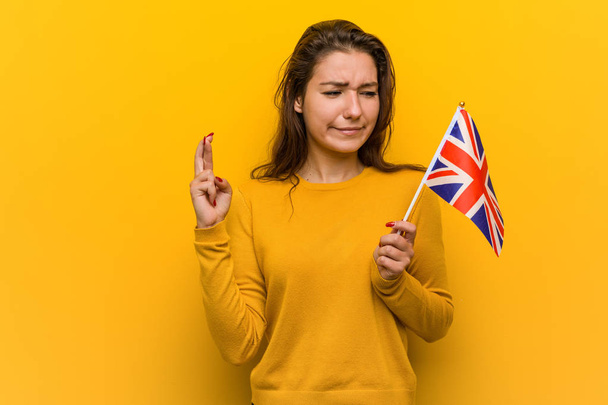 Joven mujer europea sosteniendo una bandera del reino unido cruzando dedos para tener suerte
 - Foto, Imagen
