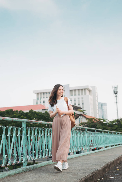 Beautiful Asian female college student holding her books stands and dresses up in street fashion cloth at outdoor public space with modern building background - Photo, image