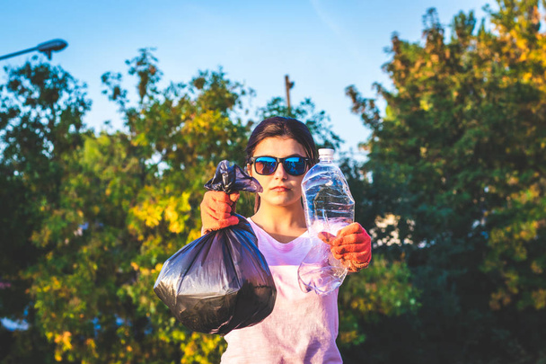 Молодая женщина держит полный мешок для мусора и использует пластиковую бутылку в руках. Концепция пластиковых и пищевых отходов. Чистая планета Земля, собирать мусор, избегать загрязнения, сохранить окружающую среду
. - Фото, изображение