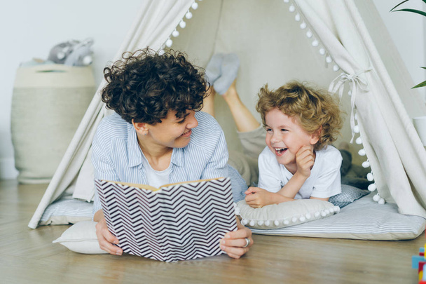 Любящая мать читает книгу симпатичному ребенку, лежащему на полу в уютной палатке в помещении
 - Фото, изображение