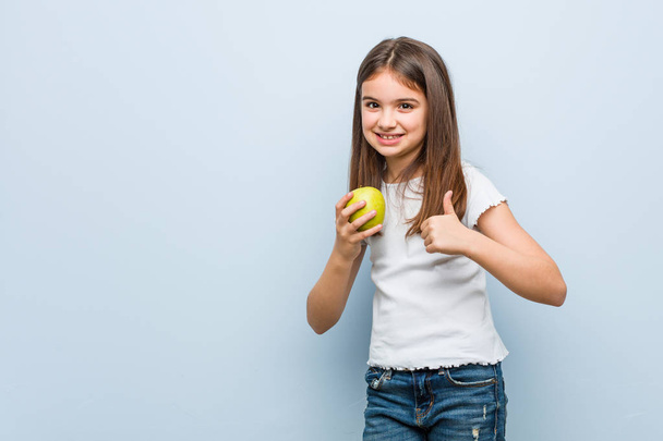 Little caucasian girl holding a green apple smiling and raising thumb up - Фото, изображение