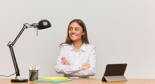 Jovem estudante mulher trabalhando em sua mesa sorrindo confiante e cruzando os braços, olhando para cima
 - Foto, Imagem