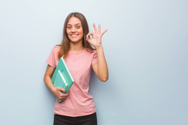 Young pretty caucasian woman cheerful and confident doing ok gesture. She is holding books. - Φωτογραφία, εικόνα
