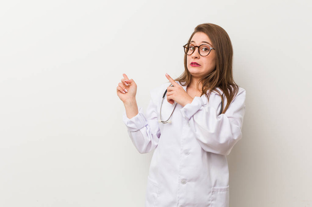 Young doctor woman against a white wall shocked pointing with index fingers to a copy space. - Photo, Image
