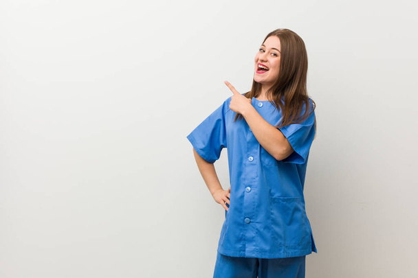 Enfermera joven contra una pared blanca sonriendo y señalando a un lado, mostrando algo en el espacio en blanco
. - Foto, Imagen