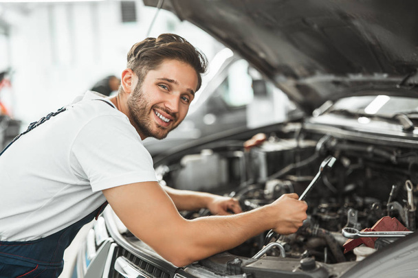 mladý usměvavý mechanik v uniformě upevnění motorových problémů v kapotě auta pracuje v servisním středisku - Fotografie, Obrázek