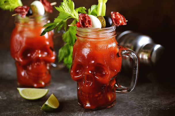 Bloody Mary cocktail in glass skull with celery sticks, pink salt, lime and canapes from canned vegetables. Halloween drink.  - Photo, Image