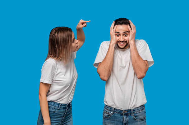 Couple of a young blond woman and brunet bearded man with mustaches in white t-shirts and blue jeans quarreling standing isolated over blue background. Concept of relationship crisis.  - Photo, Image