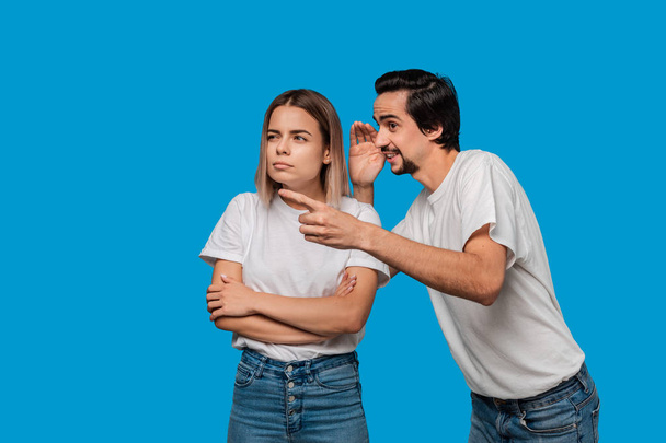 Bearded man with mustaches in white t-shirt tells secret to a young blond girl in white t-shirts and blue jeans standing isolated over blue background. Concept of secret telling. - Photo, image