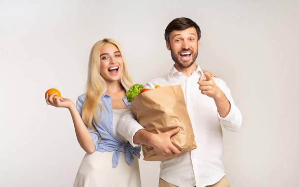 Emocionada pareja señalando el dedo en la cámara sosteniendo la bolsa de comestibles, estudio
 - Foto, imagen