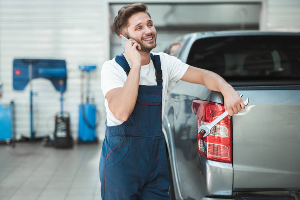 young handsome mechanic wearing uniform leaning on pickup trunk talking on the phone in car service center - Zdjęcie, obraz