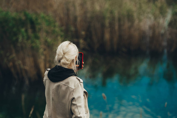 Rückansicht einer Frau mit blonden Haaren, die mit ihrem Handy ein Foto vom blauen See macht. Nahaufnahme. - Foto, Bild