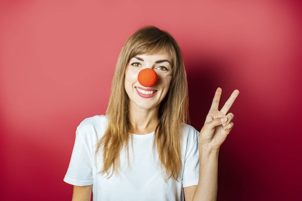 Забавная молодая женщина с красным носом клоуна на розовом фоне. День красного носа, праздник, вечеринка, облачение
 - Фото, изображение