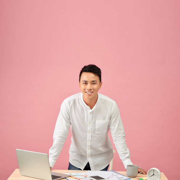Portrait of a smiling young Asian designer working on a laptop while leaning on his desk - Photo, image