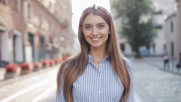 Portrait of Young Happy Charming Girl with Straight Brown Hair and Blue Eyes Wearing in Striped Dress and Fashionable Headband Smiling Looking to Camera Standing at the City Street Zoom. - Felvétel, videó