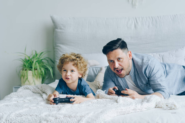 Father and child playing video game on bed at home pressing buttons on joystick - 写真・画像
