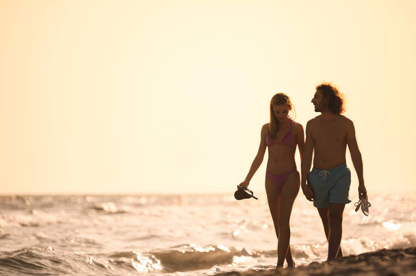 Young woman in bikini and her boyfriend walking on beach at sunset. Lovely couple - Foto, immagini