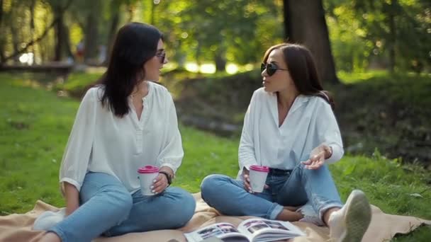 zwei stylisch glücklich lächelnde Mädchen in weißen Hemden sitzen und lachen im Park vor einem weichen Hintergrund grünen Laubs. Freundinnen haben Spaß, reden, trinken Kaffee in Pappbechern und checken ein. - Filmmaterial, Video