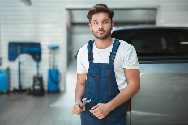 młody przystojny mechanik w mundurze gospodarstwa klucz stoi w centrum usług samochodowych - Zdjęcie, obraz