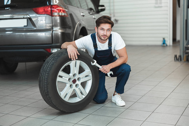 young handsome mechanic wearing uniform working in car service department fixing flat tire looks pleased - Foto, Imagen