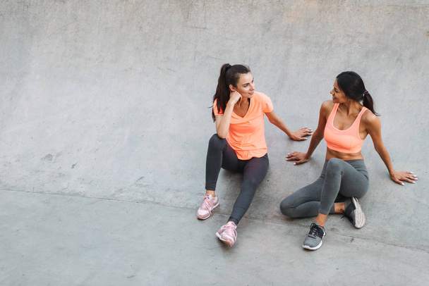 Image of attractive fitness women in sportswear talking together while sitting on concrete sports ground - Photo, Image