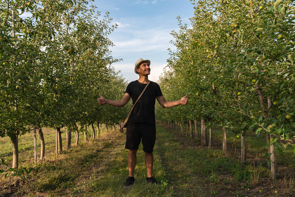 Happy young man in hat and black t-shirt standing among apples garden with wide hands and looking at up right corner - Photo, Image