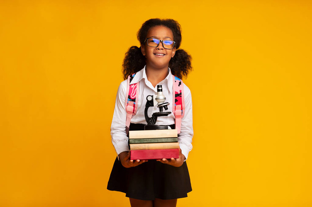 Black elementary student girl holding microscope and books, studio shot - Foto, imagen