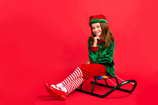 Portrait of her she nice attractive charming cheerful cheery funny sweet pre-teen elf santa claus apparel look outfit sitting on sled isolated over bright vivid shine red background - Zdjęcie, obraz