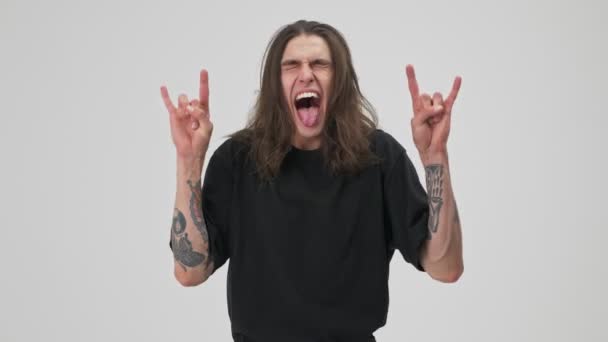 Cheerful crazy young tattooed brunette man with long hair in black t-shirt screaming and making rocker gesture with hands while looking at the camera over gray background isolated - Кадры, видео