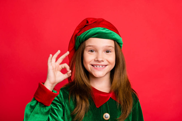 Close up photo of charming elf kid in green hat with long red head haircut showing ok sign recomme3nd eve noel event wearing fairytale cap costume isolated over red background - Foto, Imagen