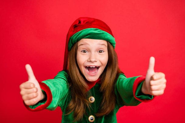 Close up photo of cheerful elf kid in hat cap having long red head showing her thumb up screaming recommend noel event wearing green costume isolated over red background - Valokuva, kuva