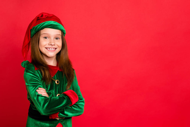 Portrait of cheerful elf kid with her hands crossed smiling ready to help in christmas night wearing green hat fantasy costume isolated over red background - Photo, image