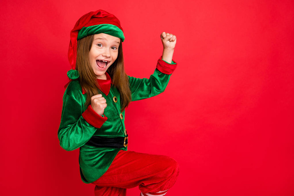 Portrait of ecstatic elf in green cap hat with long red head hairstyle scream wow omg raise fists celebrate new year partywear costume isolated over red background - Zdjęcie, obraz