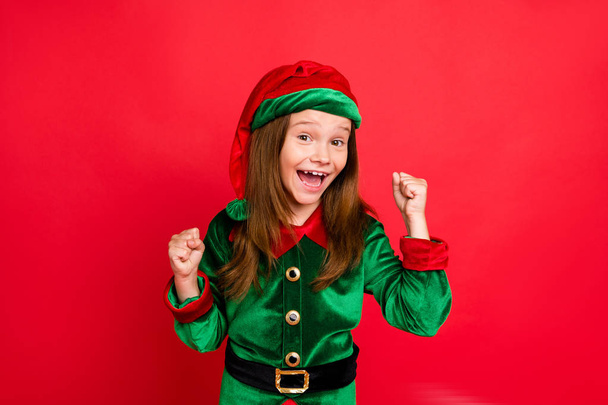 Portrait of shocked elf kid in green hat cap with long red head hairstyle raising her fists celbrate newyear party wearing fairy costume isolated over red background - Фото, изображение
