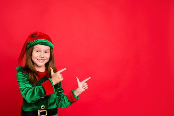 Porträt der fröhlichen Elfe mit grüner Hutmütze, die Werbung mit dem Zeigefinger zeigt, empfahl den Abend im Kostüm vor rotem Hintergrund - Foto, Bild