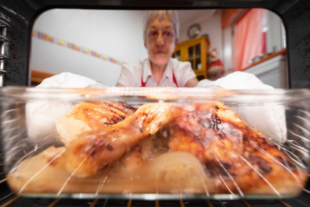 Mujer mayor tomando pollo asado del horno, visto desde el interior
. - Foto, imagen