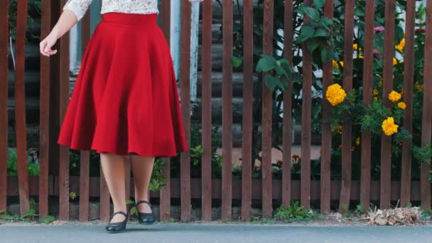 村の通りのフェンスの前で踊る長い赤いスカートの女性 - 彼女の足を踏みつぶす - 映像、動画