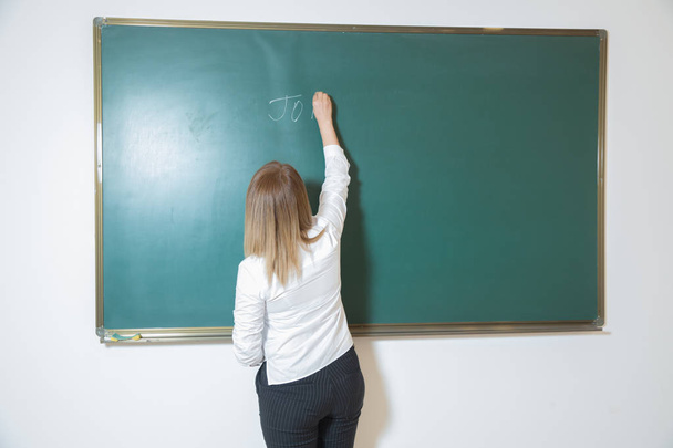 Mladá žena píše něco na tabuli. Zadní pohled na studenta nebo učitele s dlouhým brunetým účesu na prázdné zelené tabuli nebo tabuli s copyspace . - Fotografie, Obrázek