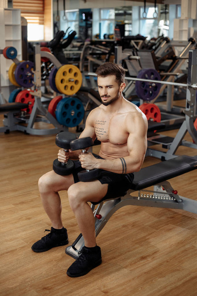 Muscular athletic young man bodybuilder fitness model focusing on exercise with dumbbell in the gym. - 写真・画像