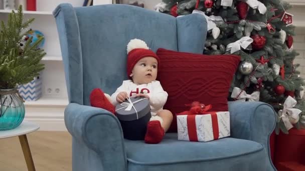 Attractive picture of sweet little baby girl in christmas clothes sitting with new year gifts in comfortable chair near christmas tree - Imágenes, Vídeo