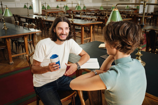 Young attractive smiling male student happily talking with friend during study break in library of university - Foto, Bild
