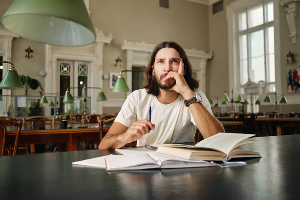 Fiatal vágyakos férfi hallgató készül vizsga elgondolkodva tanul könyvekkel könyvtárban az egyetemi - Fotó, kép